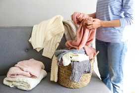 Woman sorting through bunch of sweaters of different material and knitting pattern in pile on gray sofa.